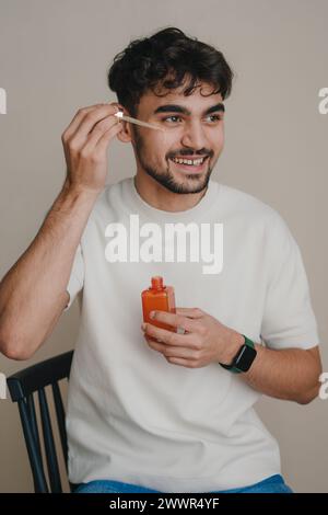 Smiling man sitting on chair against white background, looking away applying care product with pipette to area under eyes. Concept of beauty Stock Photo