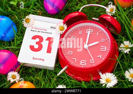 Augsburg, Bavaria, Germany - March 25, 2024: Symbolic image of the time change from winter time to summer time at Easter. An alarm clock in a meadow together with colorful Easter eggs and a calendar with the date 31 March, Easter Sunday *** Symbolbild Zeitumstellung von Winterzeit auf Sommerzeit an Ostern. Ein Wecker in einer Wiese zusammen mit bunten Ostereiern und einem Kalender mit Datum 31 März, Ostersonntag Stock Photo