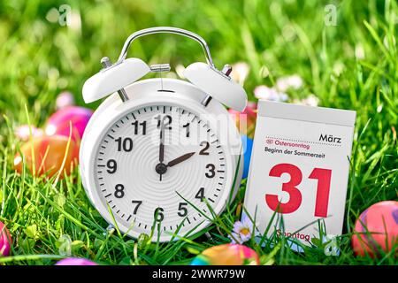 Augsburg, Bavaria, Germany - March 25, 2024: Symbolic image of the time change from winter time to summer time at Easter. An alarm clock in a meadow together with colorful Easter eggs and a calendar with the date 31 March, Easter Sunday *** Symbolbild Zeitumstellung von Winterzeit auf Sommerzeit an Ostern. Ein Wecker in einer Wiese zusammen mit bunten Ostereiern und einem Kalender mit Datum 31 März, Ostersonntag Stock Photo