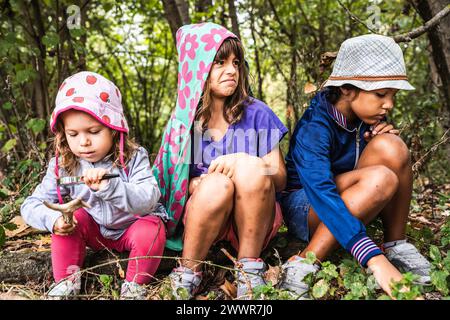 Adventurer little girls in casual clothes camping in the wild forest - One of them looking with a magnifying glass at a roe deer horn, the others maki Stock Photo