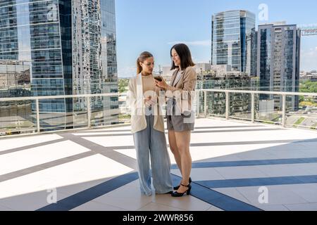 Two businesswomen standing on a terrace, one of them holding a cell phone and showing an email to her colleague. The scene is set in a city with tall Stock Photo