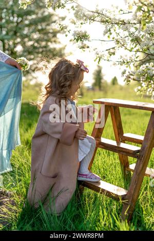 A stylishly dressed little girl with curled hair climbs a wooden ladder near a blooming pear tree Stock Photo