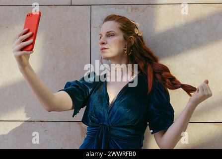 Stylish woman with red hair taking a selfie, wearing an elegant blue velvet dress complemented by striking gold jewelry. Stock Photo