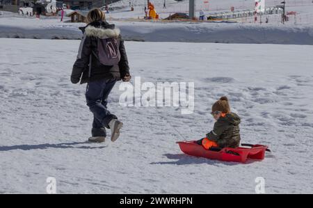 A toddler sitting on a plastic sledge is pulled along by a woman over the frozen lake at the French Alpine ski-resort of Tignes. Stock Photo