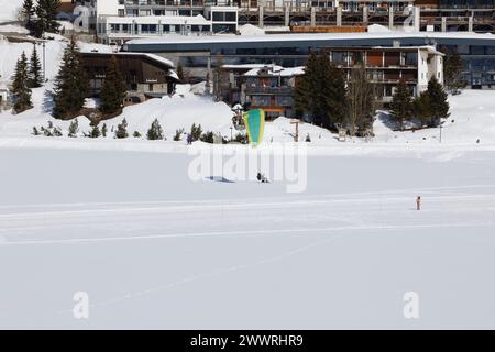 A paraglider tandem lands in front of buldings on the snow-covered, frozen, natural, lake in Tignes, a high-altitude ski-resort in the French Alps. Stock Photo