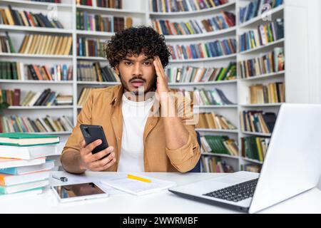 Overwhelmed islamic man sitting at desk and holding portable smartphone in personal cabinet with designer bookshelves on background. Workplace with wireless laptop, digital tablet and papers. Stock Photo