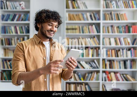 Smiling guy with curly black hair holding and looking at digital tablet in front of shelf filled with various books. Adorable hispanic male interacting with modern gadget indoors. Stock Photo