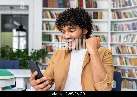 Shocked indian man working at desk surrounded by books shelves and technology. Surprised male freelancer holding smartphone in hand and appearing to reading positive news indoors. Stock Photo