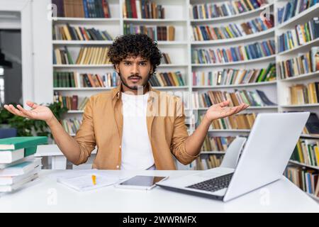 Surprised muslim man sitting at desk with modern laptop and papers surrounded by bookshelves. Male wearing white shirt and light brown jacket gesturing with both hands raised and palms up indoors. Stock Photo