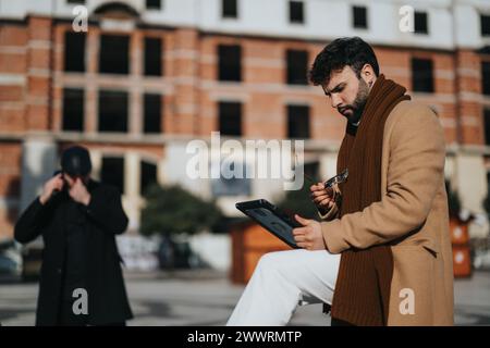 Focused businessman using a tablet while working outside. Another professional in background on phone. Urban construction site backdrop. Stock Photo