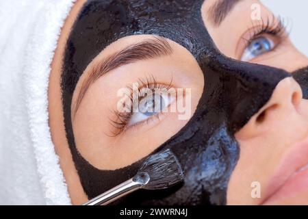 The procedure for applying a black mask to the face of a beautiful woman. Spa treatments and care of the cats in the beauty salon. Stock Photo