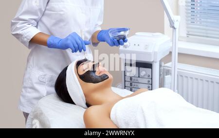 The procedure for applying a black mask to the face of a beautiful woman. Spa treatments and care in the beauty salon. Stock Photo