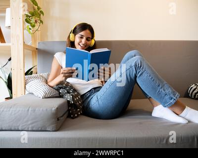 Carefree young woman reading a book on the sofa in an apartment. Literature and reading Stock Photo
