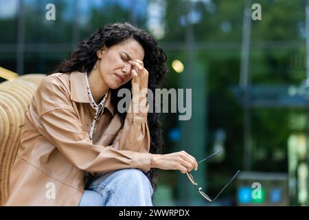 Sorrowful woman with luxurious black curly hair holding pair of glasses while touching face in nasal area. Despondent female sitting on outdoors bench and resting after hard work day on fresh air. Stock Photo