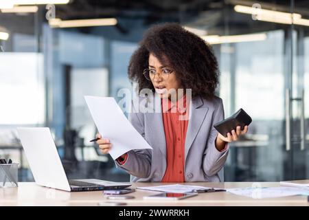 Angry female accountant holding document and calculator in hand while working in office settings. Shocked black woman in glasses sitting in front of modern laptop and performing duties indoors. Stock Photo