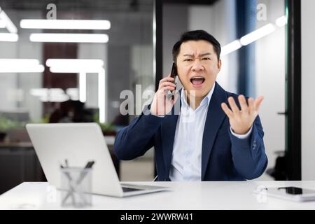 Angry chinese man screaming while having conversation on wireless smartphone on background of glassy office wall. Evil businessman using modern gadgets to staying in touch with colleagues. Stock Photo