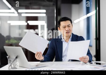 Punchy man holding documents while sitting at white desk in office environment. Handsome asian male doing work on background on glassy wall of business center. Concept of technology and people. Stock Photo