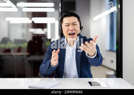 View from laptop camera of handsome asian male shouting in microphone of wireless headset while having online video call through modern gadgets. Concept of technology and business. Stock Photo