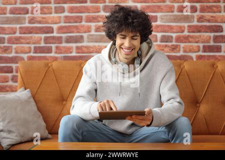 Guy sitting on a sofa and using a digital tablet with brick wall as a background Stock Photo