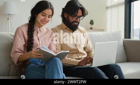 Diverse couple on couch with laptop pay bills loan payments planning household budget arabian woman wife read notes in notebook indian man husband Stock Photo