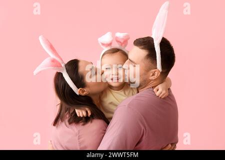 Happy parents in Easter bunny ears kissing daughter on cheeks against pink background Stock Photo