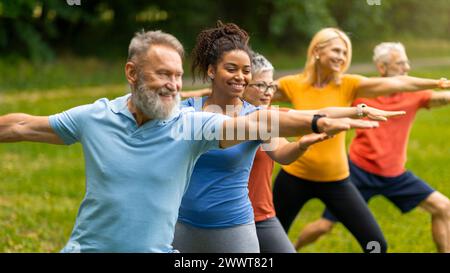 Group of diverse seniors led by instructor practicing warrior yoga pose outdoors Stock Photo