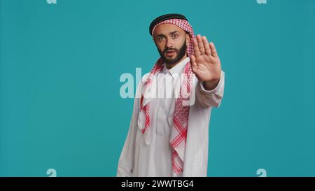 Middle eastern person does stop sign with palm up, expressing refusal and denial on studio camera. Young man presenting rejection symbol, being displeased and disappointed. Stock Photo