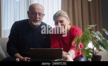 Happy senior elderly couple sit on the sofa looking at tablet computer webcam making distance video call talking. Cheerful mature elderly family onlin Stock Photo