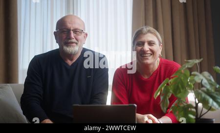 Elderly couple sitting on the couch looking into the camera, using webcam to talking with grandchildren or adult children. Webcam view, video chat and Stock Photo