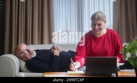 Smiling elder woman sit on sofa at home check information on document, taking notes, doing paperworks. Smiling old woman talk and work with husband wh Stock Photo