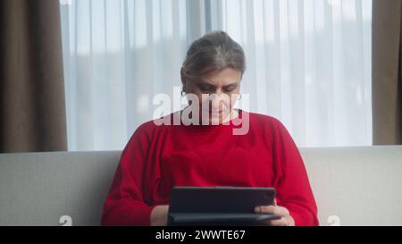 Middle aged old woman sit on sofa holding digital tablet reading e-book, enjoying using computer pad apps, browsing internet shopping in living room Stock Photo