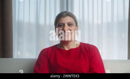Smiling mature lady sitting on sofa looking at camera at home. Happy middle aged senior woman posing in living room. Portrait medium close-up Stock Photo