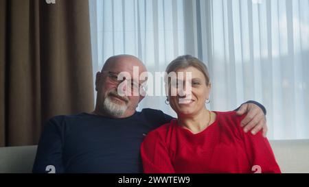 Happy elderly family couple sitting on sofa looking at the camera and smiling, man embracing his wife, retired grandfather and grandmother portrait Stock Photo
