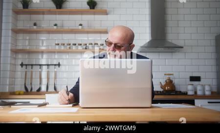 Elderly business man with glasses sit in kitchen using laptop computer, taking notes, analyze data, looking through papers busy in paperwork Stock Photo