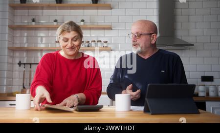 Senior parents sit in kitchen doing paperwork, checking calculating bills bank loan payment, discuss unpaid debt taxes. Old grandparents family money Stock Photo