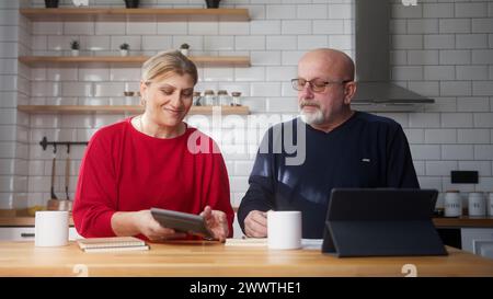 Retired married couple sit in kitchen doing paperwork, checking calculating bills bank loan payment, discuss unpaid debt taxes. Old grandparents famil Stock Photo