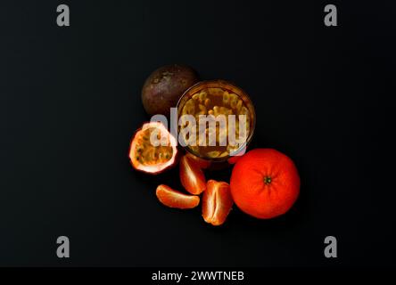 A glass of fruit juice with seeds on a black background, next to pieces of ripe mandarin passion fruit. Top view, flat lay. Stock Photo