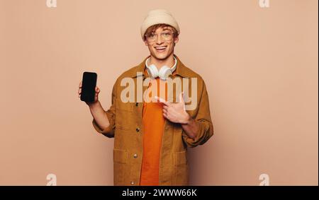 Happy young man in casual clothing stands in a vibrant studio. He holds a mobile phone, pointing and recommending an app on the touchscreen. Wearing g Stock Photo