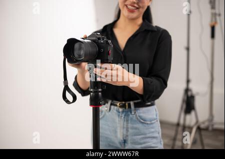 A cropped image of a professional Asian female photographer is adjusting her DSLR camera on a tripod stand, preparing shooting equipment in the studio Stock Photo