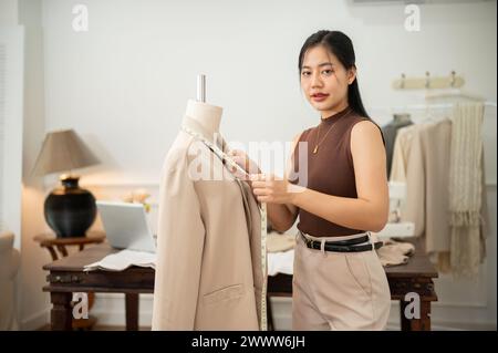 A professional and confident Asian female tailor or fashion designer is working in her studio, adjusting her clothing patterns and measuring sizes wit Stock Photo