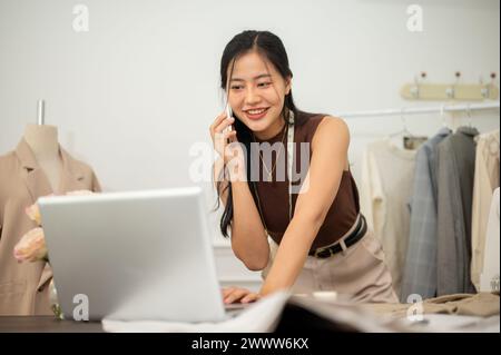 A professional, busy Asian female fashion designer or tailor is talking on the phone with her client while working on her laptop in her atelier studio Stock Photo