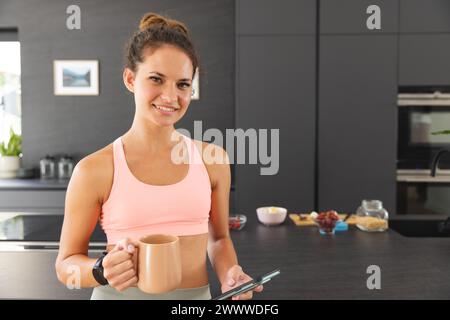 A young Caucasian woman smiles, holding a smartphone in the kitchen at home Stock Photo