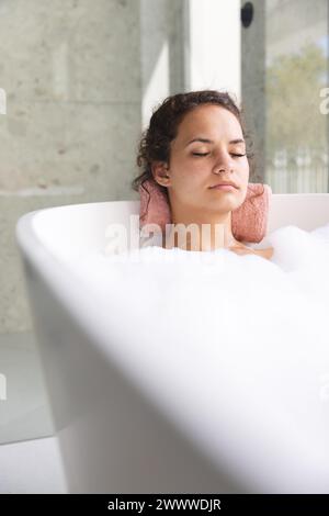 Young woman relaxes in a bubble bath at home, eyes closed in serenity, with copy space Stock Photo
