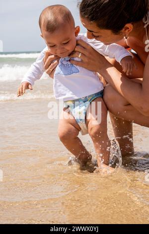 Vertical photograph of a young mother and her baby enjoying the beach for the first time, playing in the water and jumping with the waves of the sea. Stock Photo
