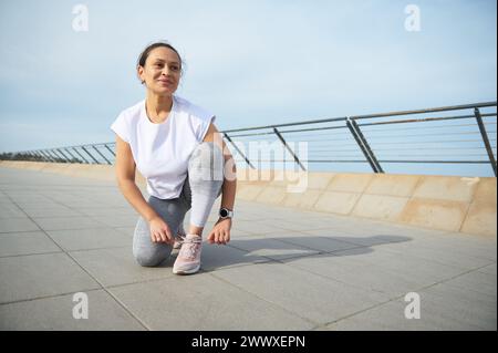 Happy multi ethnic woman runner tying laces on sports shoes, smiling looking away, getting ready for morning run on the promenade. Running nd jogging Stock Photo