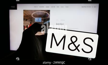 Person holding mobile phone with logo of British retail company Marks and Spencer Group plc in front of business web page. Focus on phone display. Stock Photo