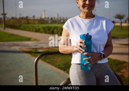 Cropped view of happy smiling fit woman in activewear holding bottle of water after fitness workout outdoors. Sportswoman holding bottle of water, rel Stock Photo