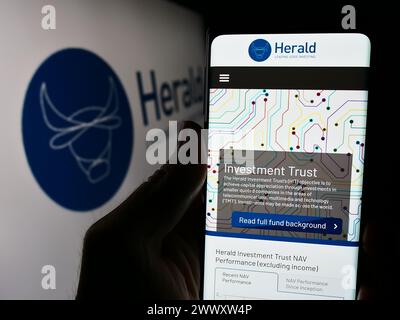 Person holding cellphone with webpage of British company Herald Investment Trust plc in front of logo. Focus on center of phone display. Stock Photo