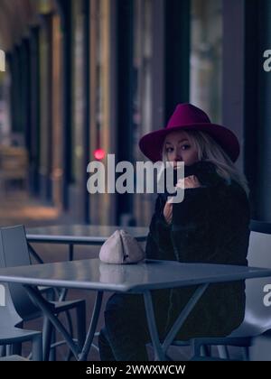 Woman in a magenta hat and black coat seated outdoors at night, city lights in the background Stock Photo