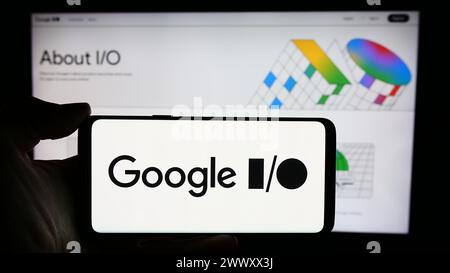 Person holding cellphone with logo of annual developer conference Google IO in front of business webpage. Focus on phone display. Stock Photo